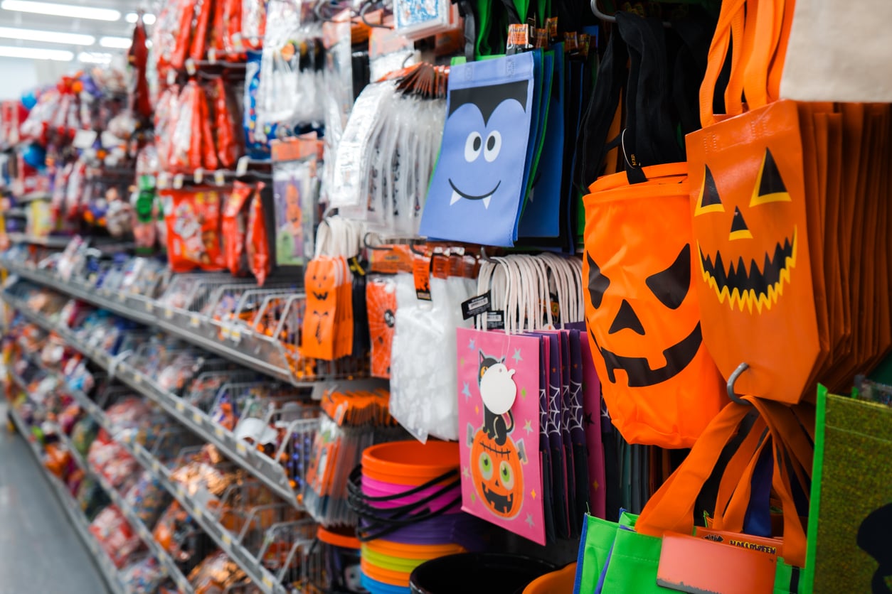 A store aisle displays colorful Halloween bags with jack-o’-lanterns and vampire faces, surrounded by various Halloween-themed items and decorations on shelves.