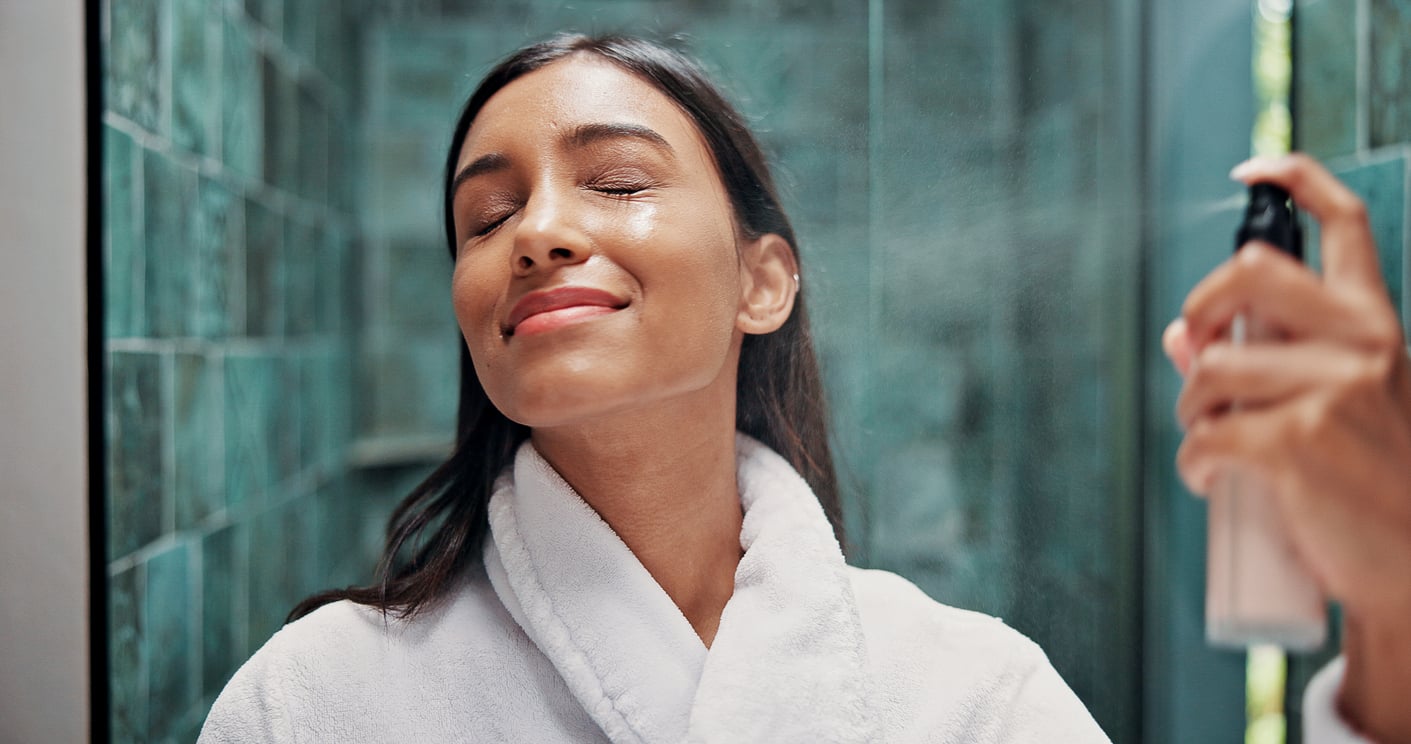 A person in a white bathrobe smiles with eyes closed while misting their face with a spray bottle in a tiled bathroom, reflecting how women are changing their spending behaviors by investing more in self-care and wellness routines.