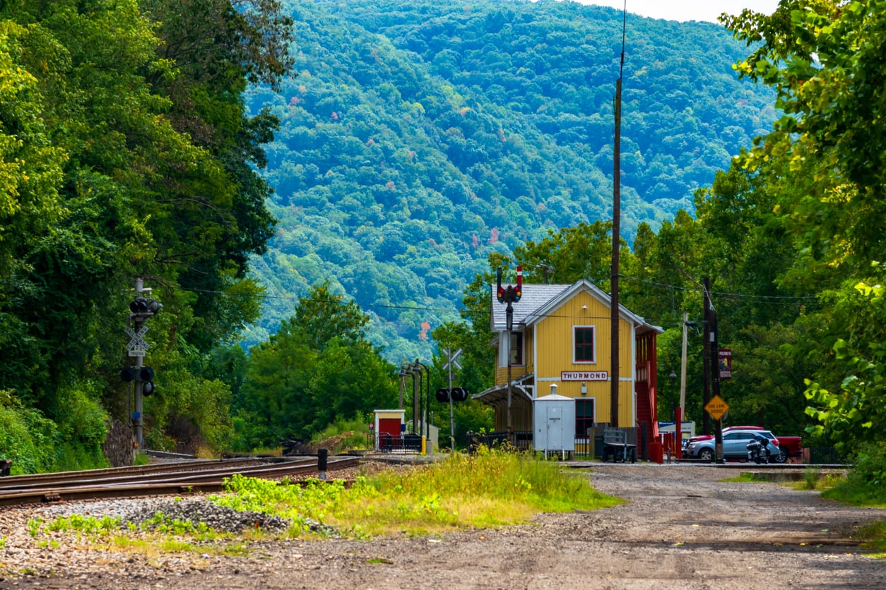 A yellow building marked “Flemington” stands beside railroad tracks, surrounded by trees and hills under a bright sky—a charming scene reminiscent of classic American towns. Signal lights, a few parked cars, and utility poles are visible near the station.
