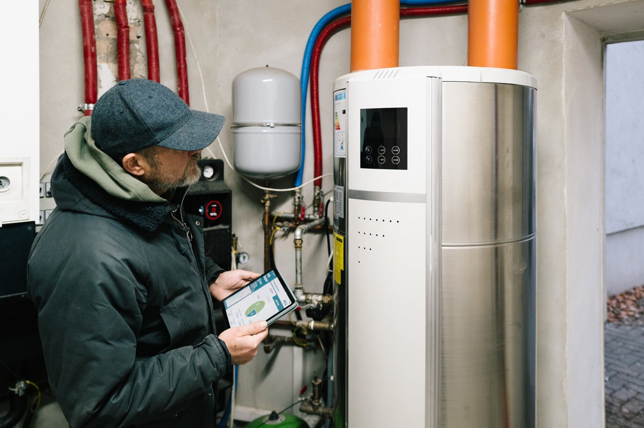 A person in a jacket and cap uses a tablet to monitor or control a modern heating system in a utility room with visible pipes and equipment.