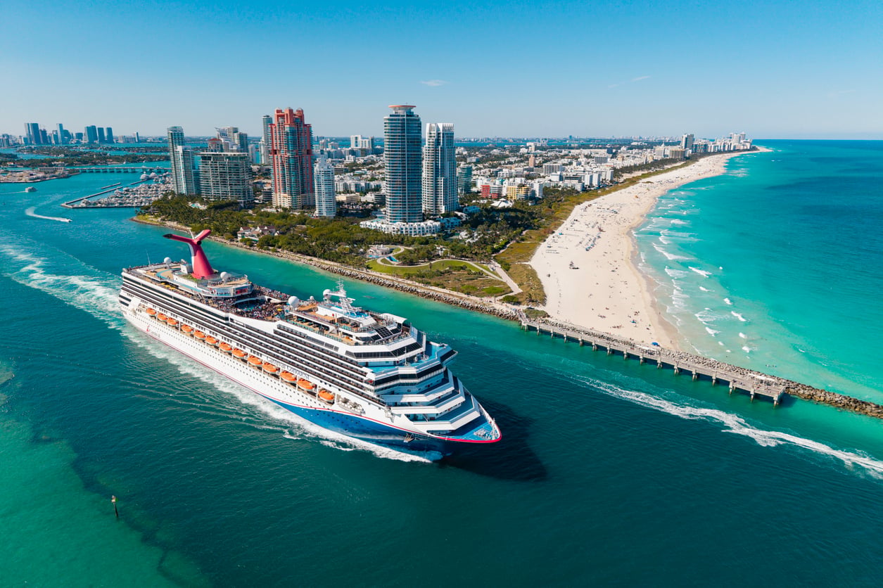 A large cruise ship sails past a sandy beach and seaside city with tall buildings under a clear blue sky, turquoise water, and a winding coastline.