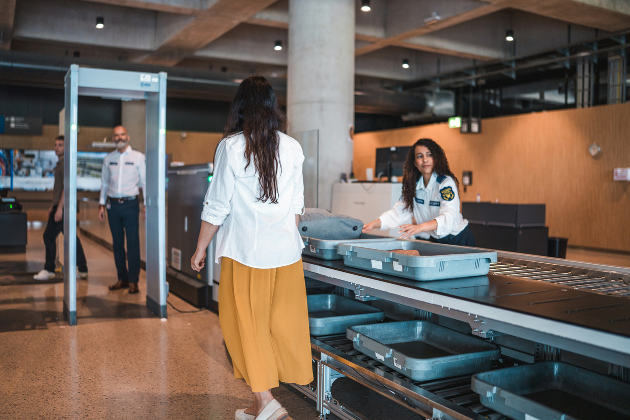 A woman walks through airport security, placing a gray sweater in a tray on the conveyor belt while a security officer observes and another person walks through a metal detector in the background.