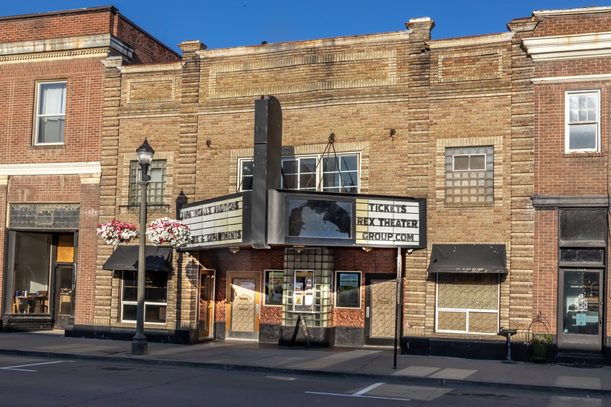 A historic brick movie theater with a vintage marquee displaying “TICKETS REX THEATER GROUP.COM.” Two flower baskets hang from a nearby streetlamp, and the empty street hints at secret movie theater hacks under a clear blue sky.
