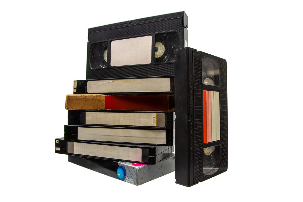 A stack of VHS tapes and cases, some standing upright and others lying flat, evokes the nostalgic entertainment choices of Middle-Class Moms in the 80s and 90s, isolated on a white background.