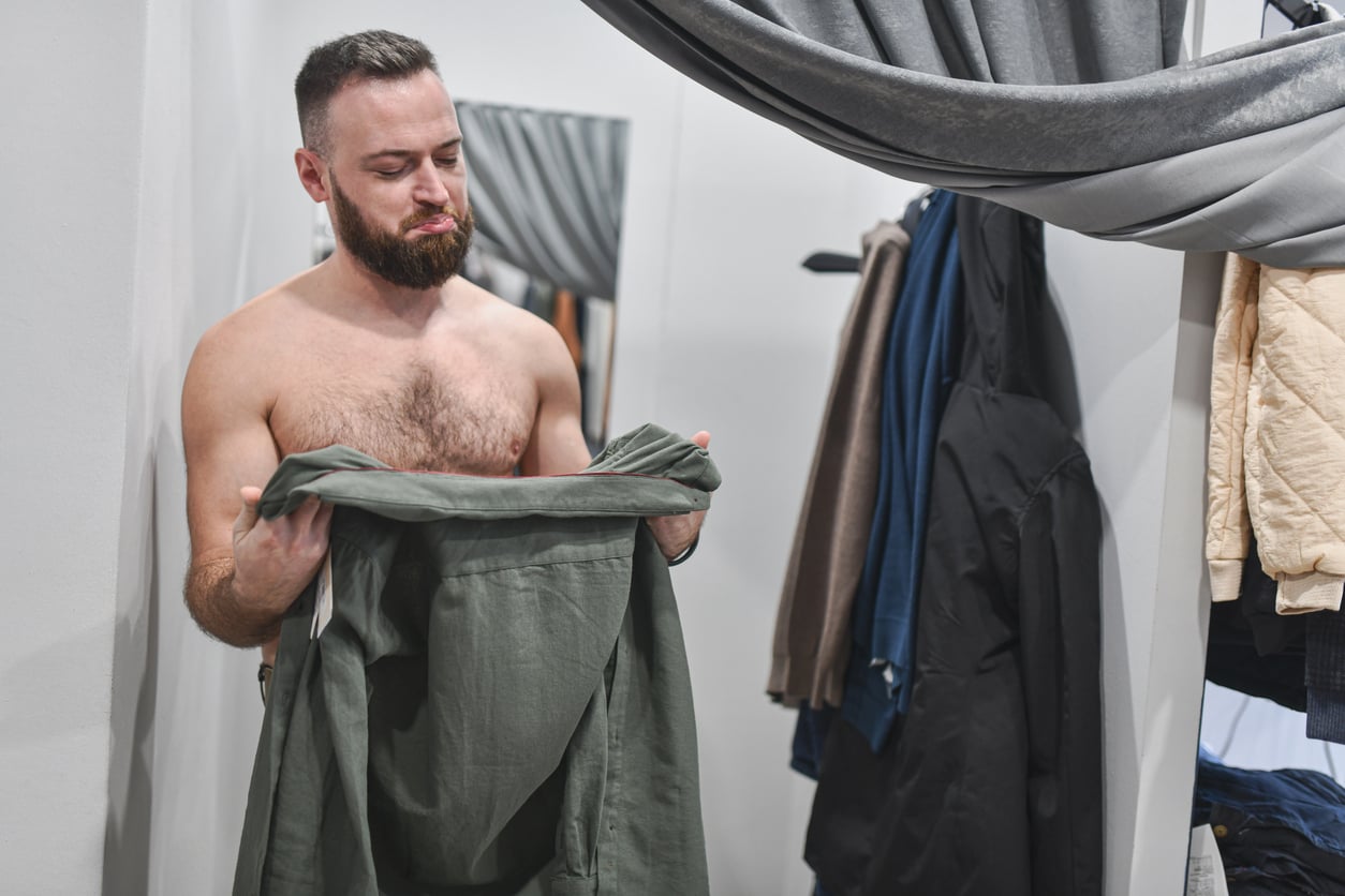 A bearded man stands shirtless in a fitting room, frowning while holding a green shirt. Practicing money saving habits, he carefully considers his purchase as various jackets and clothes hang nearby.
