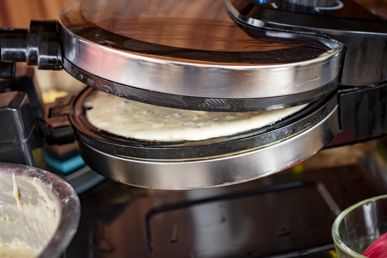 A close-up of a waffle maker, one of the must-have kitchen gadgets, with batter being cooked inside. The lid is partially closed, and the batter is visible on the hot plate. A bowl with extra batter sits in the foreground.