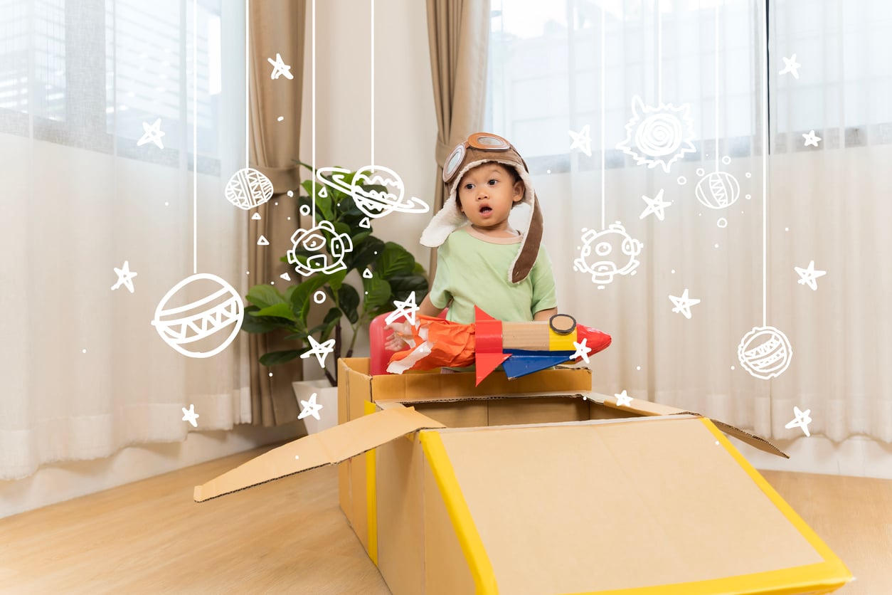 A young child wearing an aviator hat sits in a cardboard box shaped like a spaceship, surrounded by hand-drawn stars and planets, pretending to explore space in a bright living room.