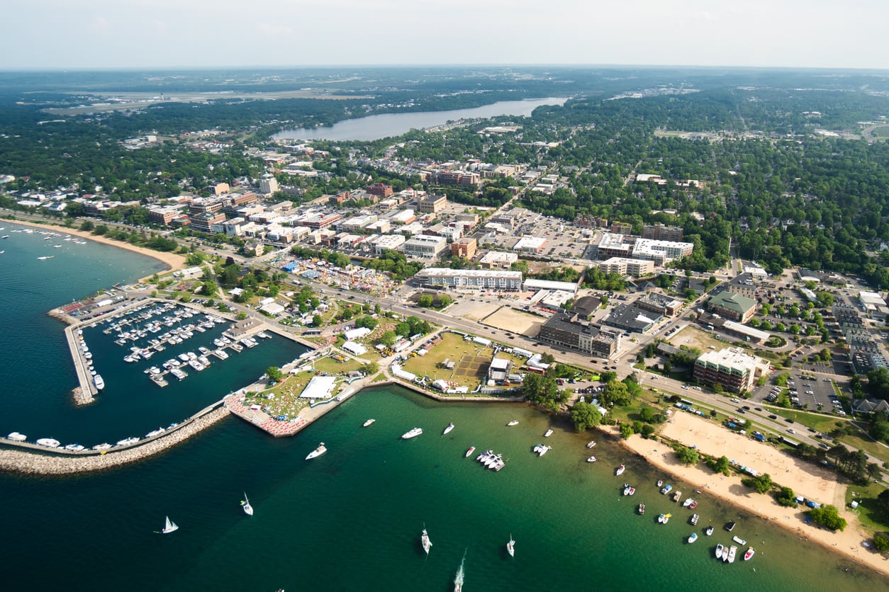 Aerial view of a lakeside town with a marina, sandy beach, green trees, boats on the water, and buildings spread throughout the area under a bright sky—one of the 20 travel destinations that perfectly blends natural beauty with vibrant waterfront living.
