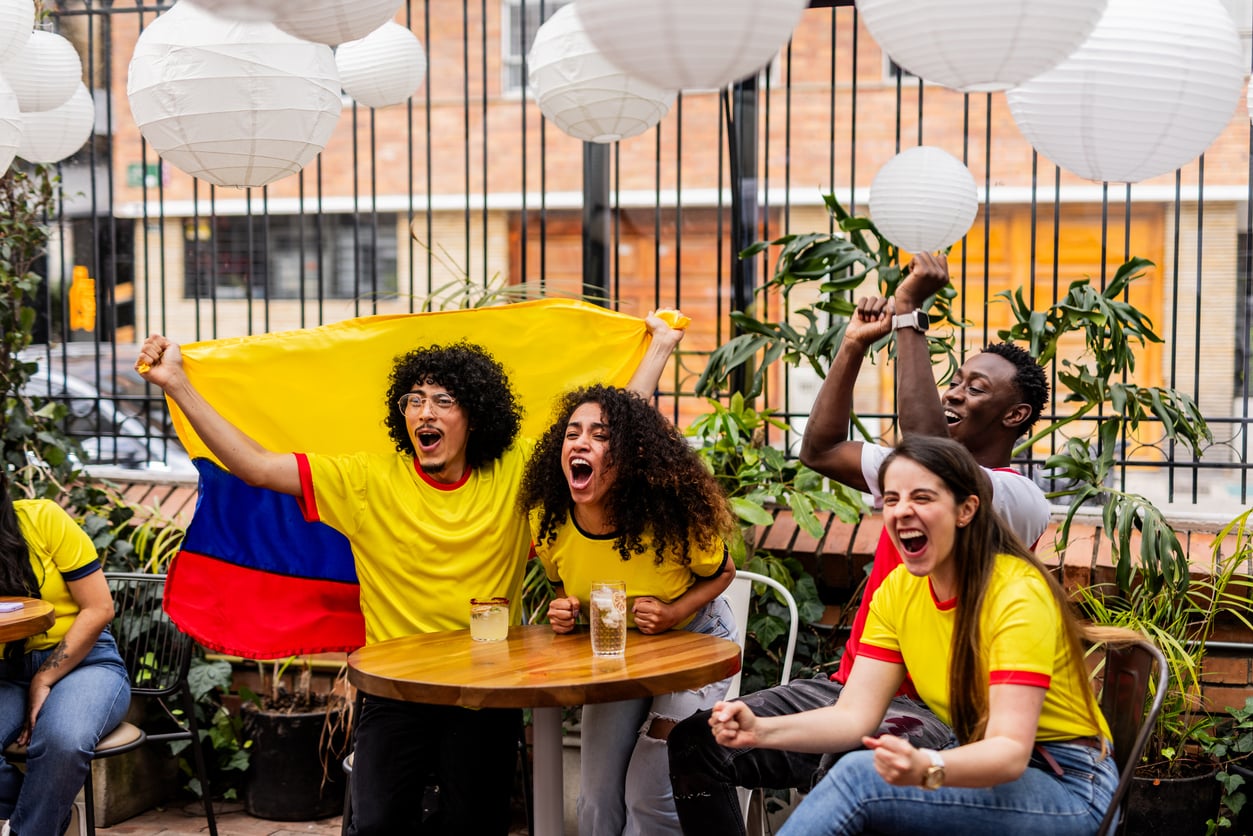 Four people in yellow shirts cheer excitedly at a table outdoors, holding a Colombian flag. White lanterns hang above, and drinks are on the table. They appear to be watching a sports event.