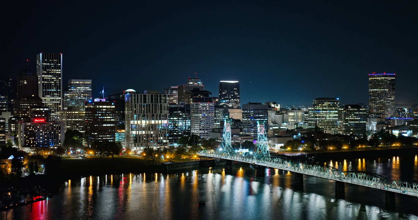 A city skyline at night features illuminated buildings and a lit bridge reflecting on the river below, with trees lining the waterfront.