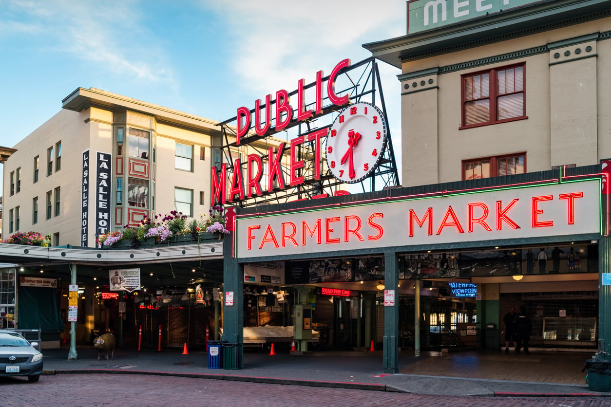 Pike Place Market entrance with large neon "Public Market" and "Farmers Market" signs, clock, flower boxes, and surrounding historic buildings on a cobblestone street in Seattle. Travel backup plans. 