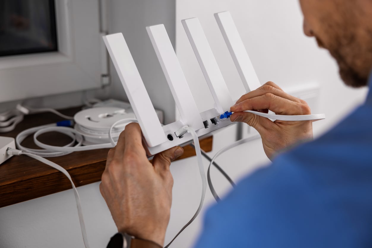A person is plugging an Ethernet cable into the back of a white wireless router with four antennas, near a window on a wooden shelf, ensuring a reliable connection to help reduce monthly bills.