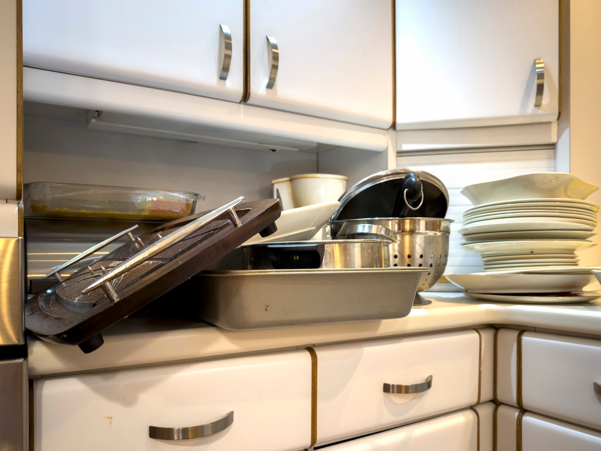 A kitchen counter cluttered with dirty dishes, pots, pans, a colander, stacked plates, and various kitchen gadgets sits in front of white cabinets.