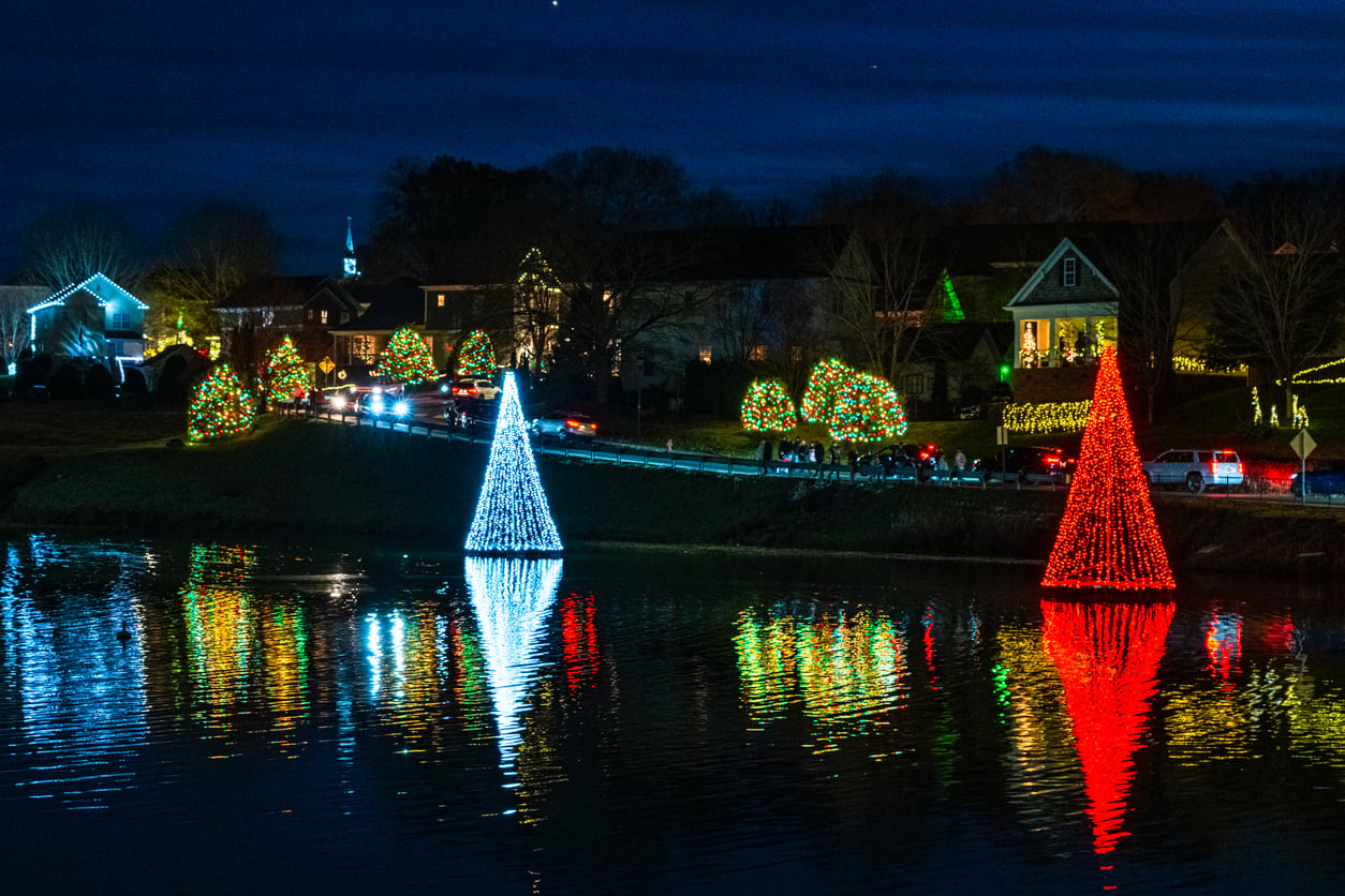 Houses and trees decorated with colorful holiday lights are reflected in a lake at night, evoking the festive charm of the best Christmas towns in America. Two cone-shaped light displays, one red and one white, stand prominently by the water.