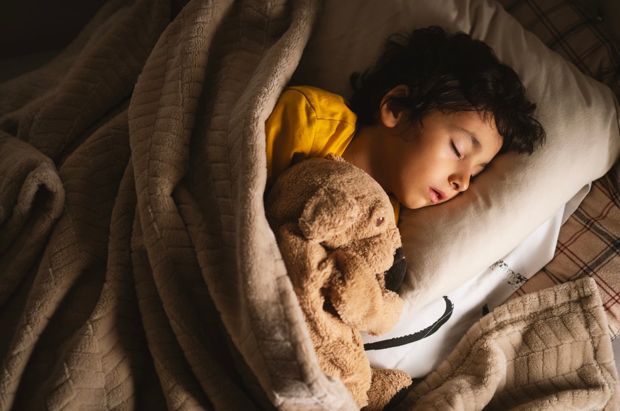 A young child with dark hair sleeps peacefully in bed, cuddling a brown stuffed animal under a beige blanket, wearing a yellow shirt.