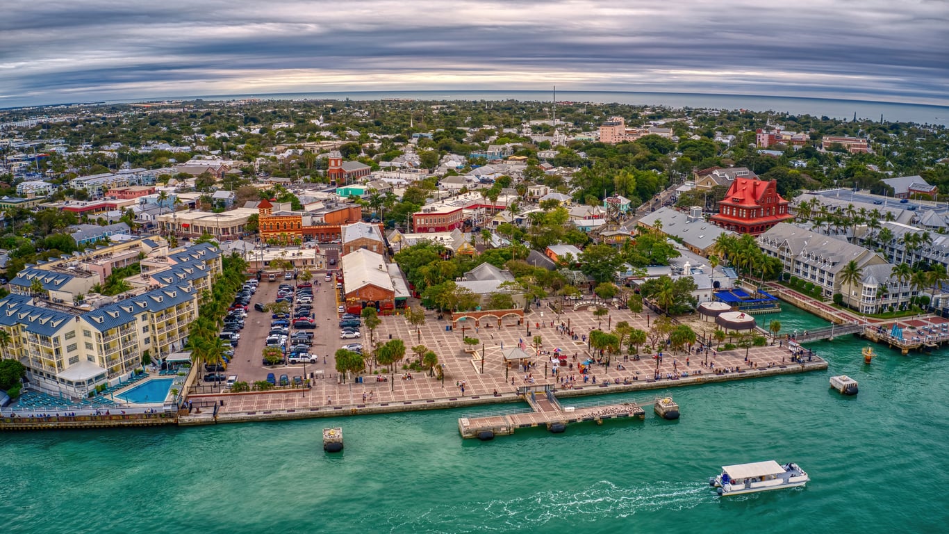 Aerial view of a coastal town with boats at a pier, turquoise water, historic buildings, and lush greenery under a cloudy sky—an idyllic scene that captures the charm of top travel destinations.