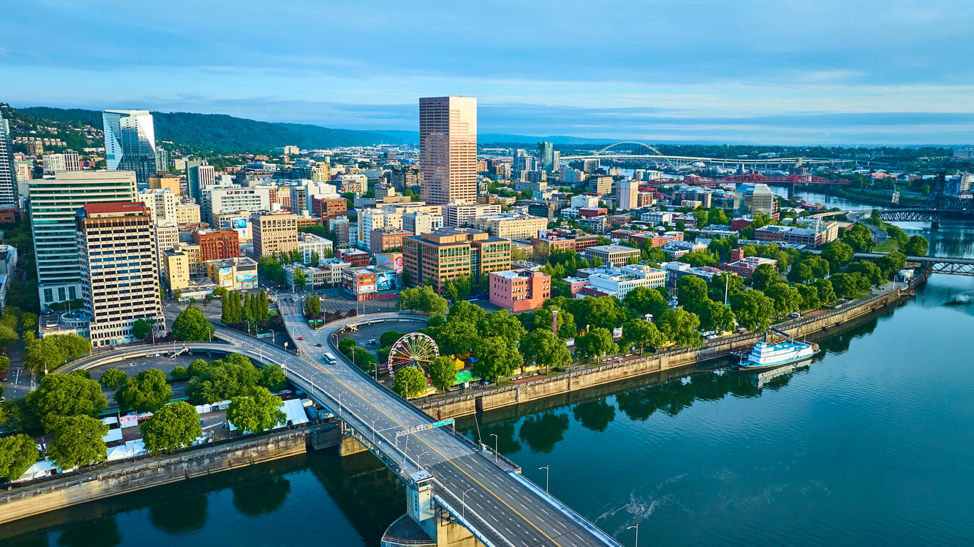 Aerial view of downtown Portland, Oregon, showing city buildings, bridges, green trees, a ferris wheel, and the riverfront beside the Willamette River on a clear day.