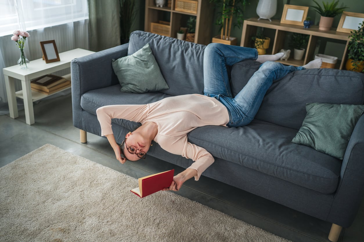 A person wearing glasses lies upside down on a gray couch, with their legs on the backrest and head hanging off the seat, holding a red book open near the floor in a cozy, well-lit living room—a perfect spot for relaxing while catching up on money saving habits.