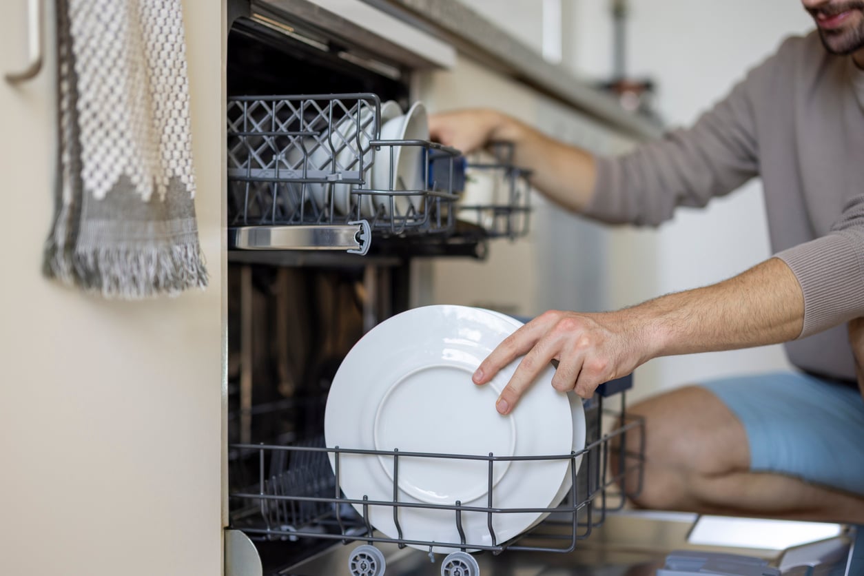 A person is loading clean plates and dishes into a dishwasher in a modern kitchen. Only their hands and part of their body are visible as they place a white plate on the bottom rack.