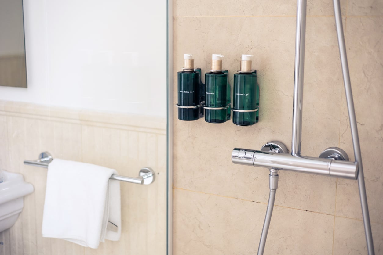 Three green soap and shampoo dispensers are mounted on a beige tiled shower wall next to a modern silver shower fixture—a common feature reflecting recent hotel changes; a white towel hangs on a rail nearby.