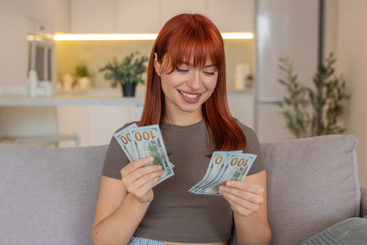 A woman with red hair sits on a sofa, smiling while holding several $100 bills in her hands. A modern kitchen is visible in the background.