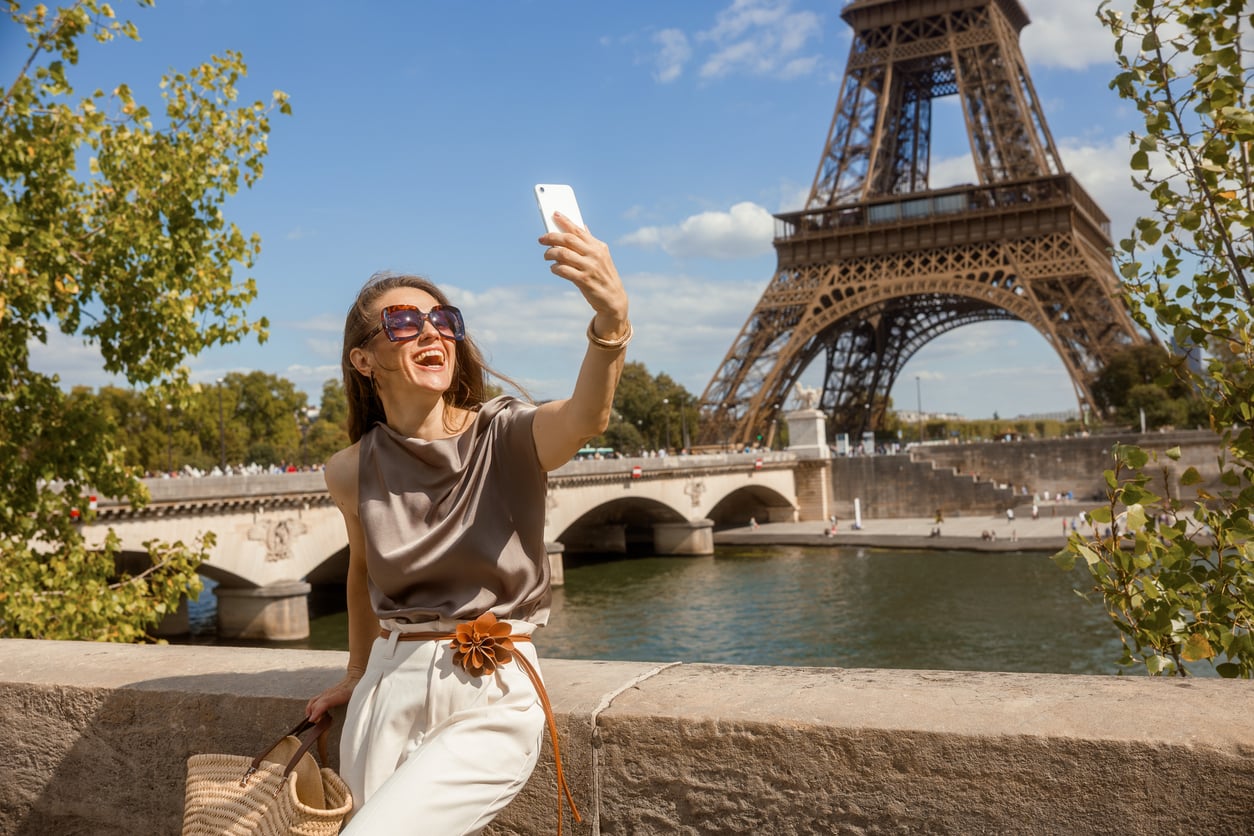 A woman wearing sunglasses takes a selfie near the Eiffel Tower on a sunny day, holding a woven bag and smiling with a river and bridge in the background.