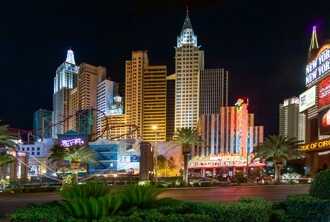The image shows the brightly lit New York-New York Hotel & Casino in Las Vegas at night, with replicas of New York skyscrapers, neon signs, palm trees, and a roller coaster visible in the scene.