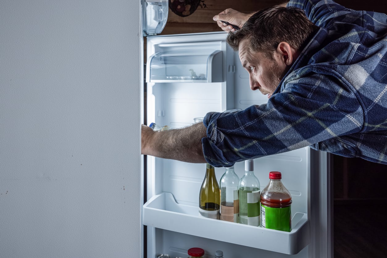 A man in a plaid shirt is looking intently inside an open refrigerator, reaching in with one hand. The fridge door has bottles and jars on the shelves. The scene is lit by the fridge’s interior light.