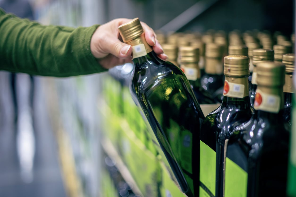 A person in a green sleeve selects a dark glass bottle with a gold cap from a shelf lined with similar bottles—once popular but now among the products people no longer buy—in a store.