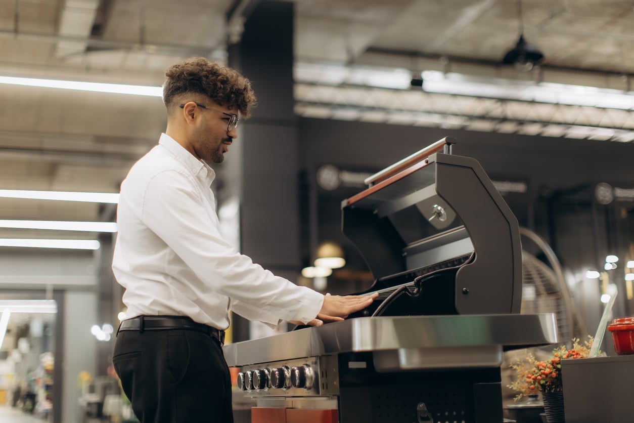 A man in a white shirt examines a gas grill in a store, lifting the lid to check the inside—one of those products people no longer buy as frequently. The background shows shelves and industrial lighting, giving the setting a modern, indoor feel.