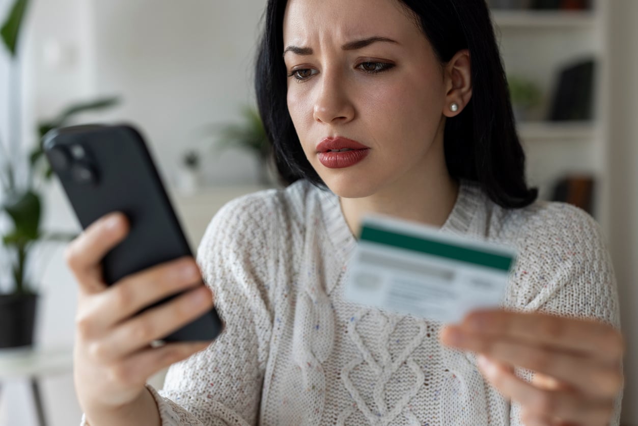 A woman with dark hair wearing a white sweater looks concerned while holding a smartphone in one hand and a bank card in the other, sitting indoors, possibly worried about managing household expenses.