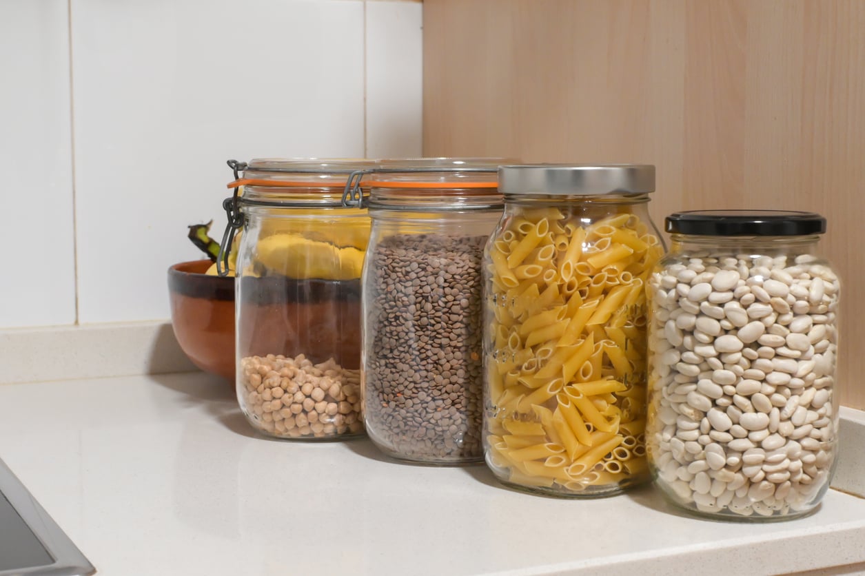 Four glass jars filled with dried chickpeas, lentils, pasta, and white beans are lined up on a kitchen countertop with a brown bowl and bananas in the background, featuring pantry staples you can find at Walmart.