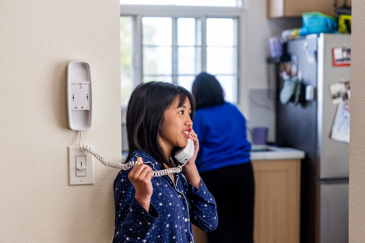 A young girl in star-patterned pajamas talks on a corded phone, standing by a wall, while an adult—capturing the essence of Middle-Class Moms in the 80s and 90s—is in the kitchen in the background near a refrigerator.