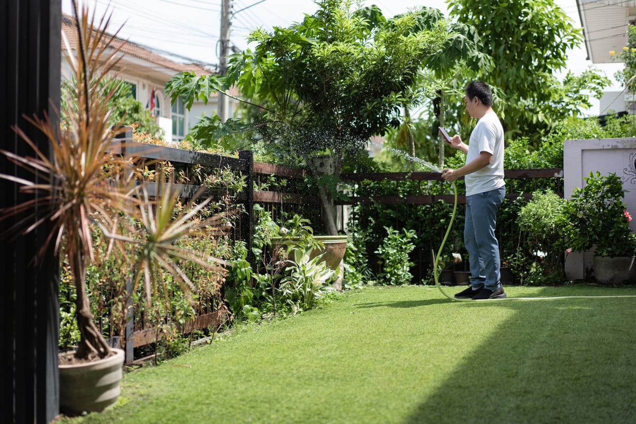 A man wearing a white shirt and jeans waters plants in a garden with a hose on a sunny day, tending to his green space while managing household expenses, surrounded by potted plants, trees, and green grass near a wooden fence.