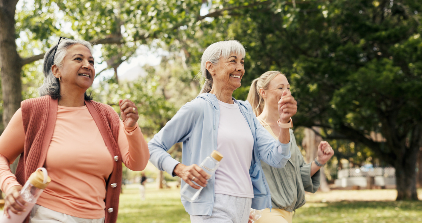 Three older women walk together in a park, smiling and holding water bottles. Dressed in casual, sporty clothing, they enjoy a sunny day outdoors—a fun, healthy way to save money while staying active amidst the green trees.