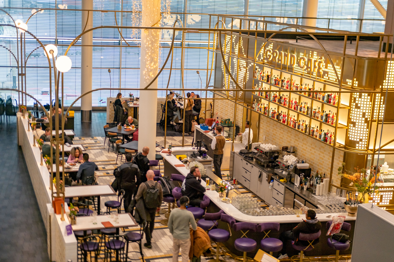 A bright, modern cafe with people sitting at tables and the bar. Shelves of colorful bottles line the back wall, while staff prepare drinks. Large windows let in natural light, creating a lively, welcoming atmosphere.