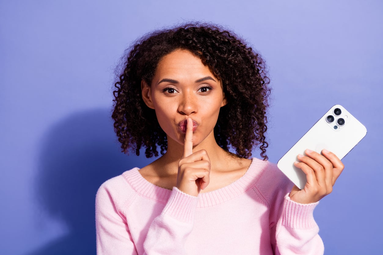 A woman with curly hair holds a smartphone in one hand and makes a shushing gesture with her finger to her lips, standing against a purple background.