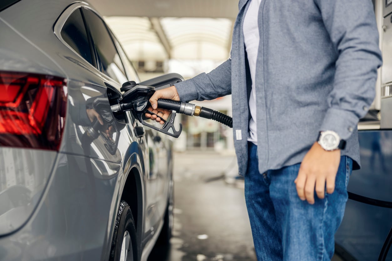 A person in a gray shirt and jeans is refueling a silver car at a gas station, holding a fuel nozzle inserted into the tank—a simple step in essential car maintenance hacks. The focus is on their hand and the fueling process.