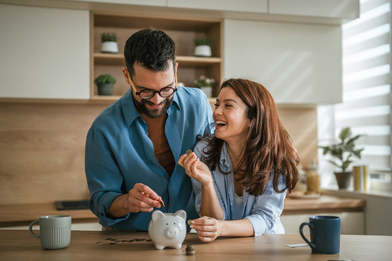 A smiling couple stands in a kitchen, happily placing coins into a piggy bank on the counter, enjoying the benefits of renting as they save together. Mugs and spare coins nearby enhance this cheerful scene of shared financial goals.