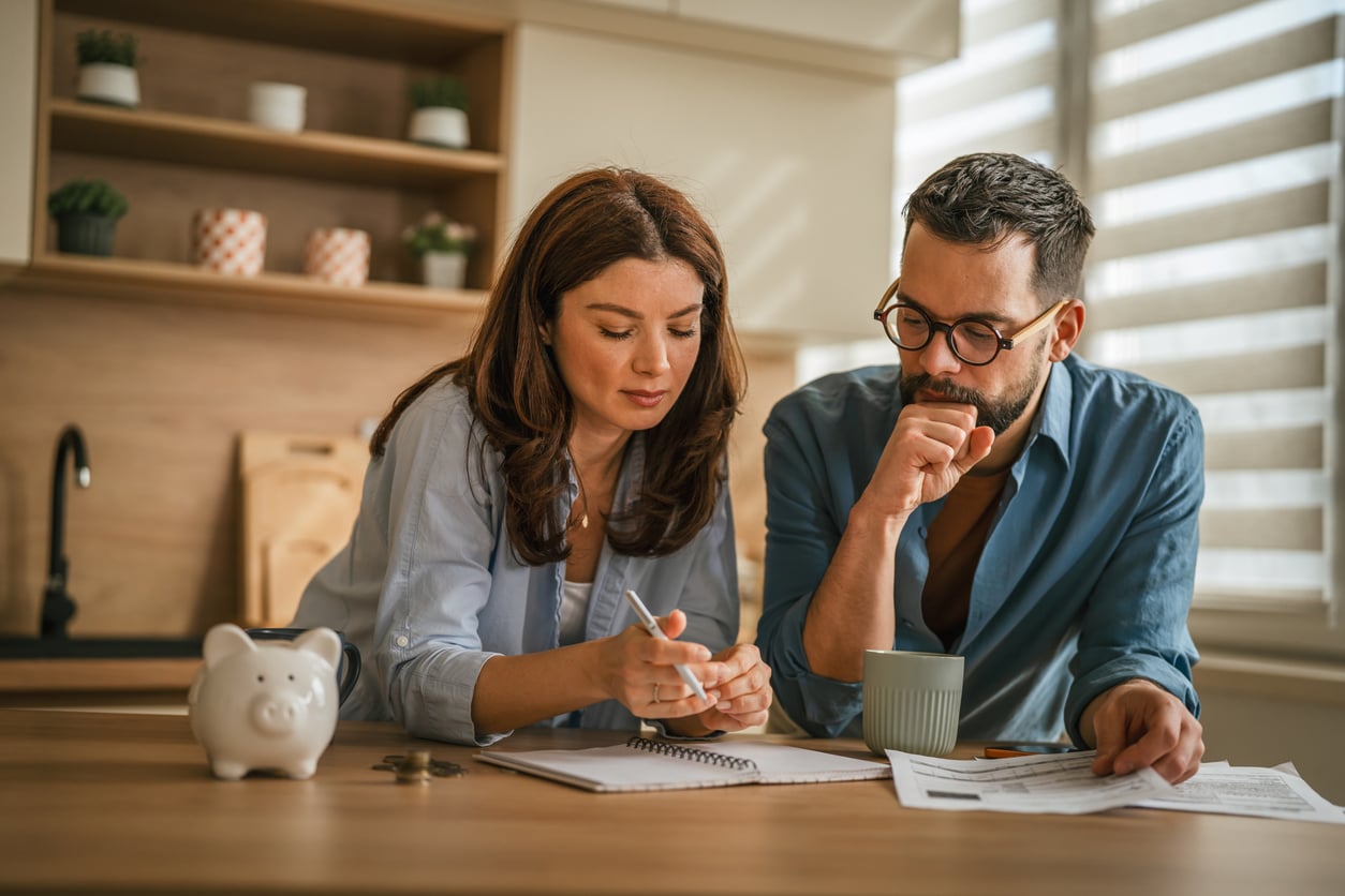 A couple sits at a kitchen table, reviewing papers and taking notes. A piggy bank and coins are on the table, suggesting they are planning their finances or budgeting together, perhaps even looking to avoid hotel booking mistakes as they organize their next trip.