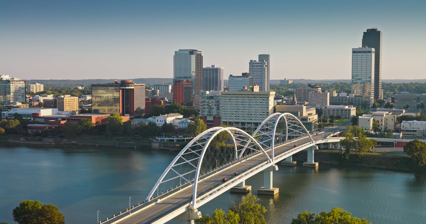 A modern, travel-friendly bridge spans a river toward a city skyline with mid-rise and high-rise buildings under a clear blue sky, surrounded by trees and water.