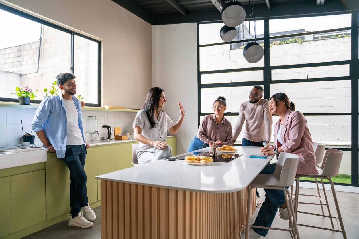 Five people in casual clothes gather and chat around a kitchen island with plates of food, smiling and enjoying each other's company in a bright, modern kitchen with large windows—perhaps sharing stories about their recent vacation swaps.