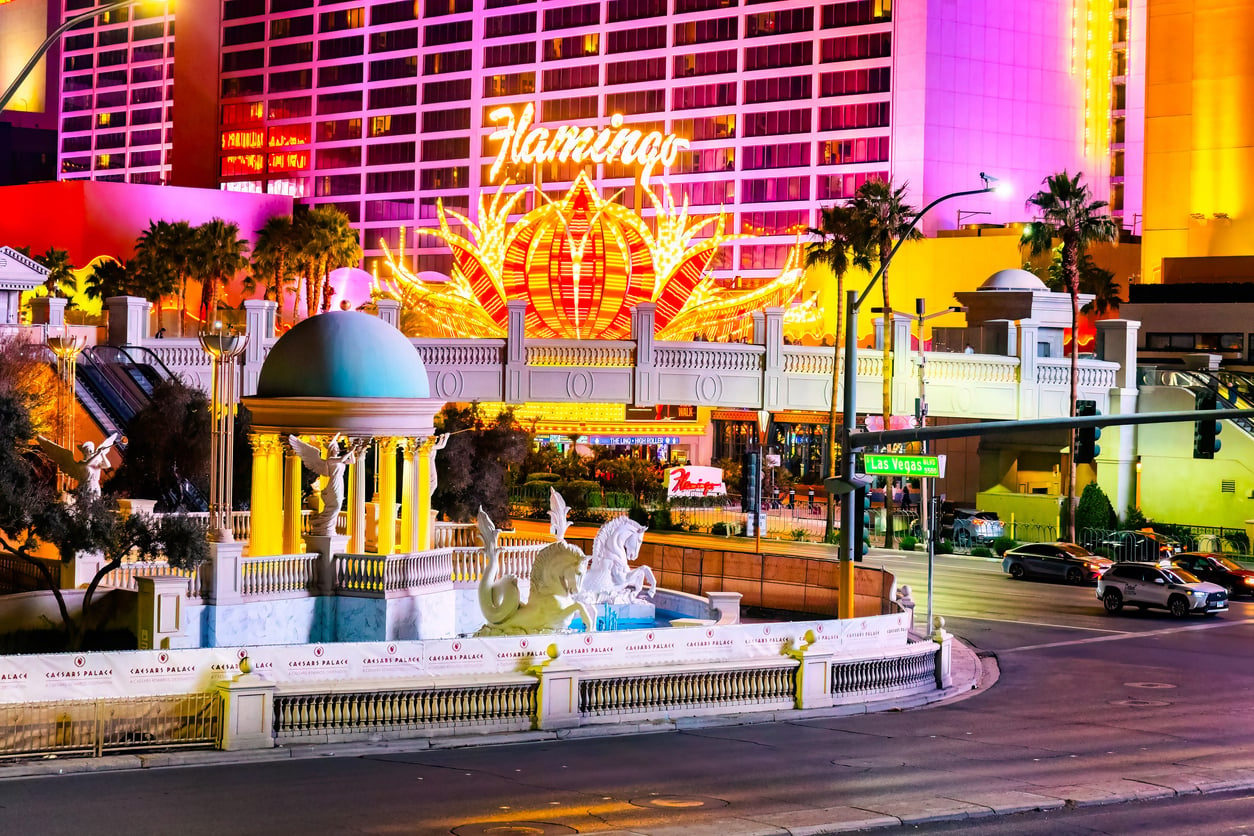 Flamingo hotel exterior at night in Las Vegas, brightly lit with neon lights. Palm trees, statues, and a fountain are visible in the foreground, with cars and people on the street nearby.