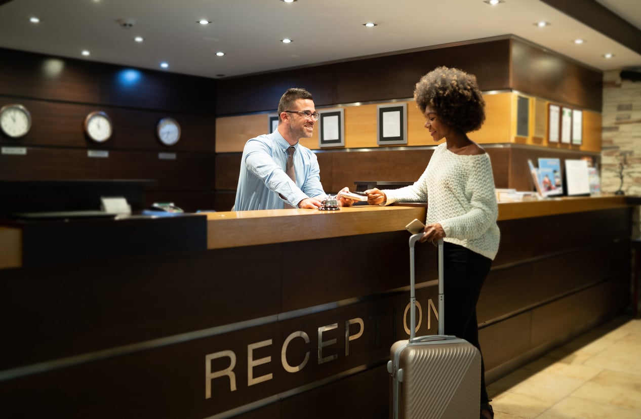 A woman with a suitcase checks in at a hotel reception, handing her ID to a smiling receptionist behind the counter. The reception area features clocks and framed documents on the wall, reminding guests of the importance of avoiding common hotel booking mistakes for a smooth check-in experience.