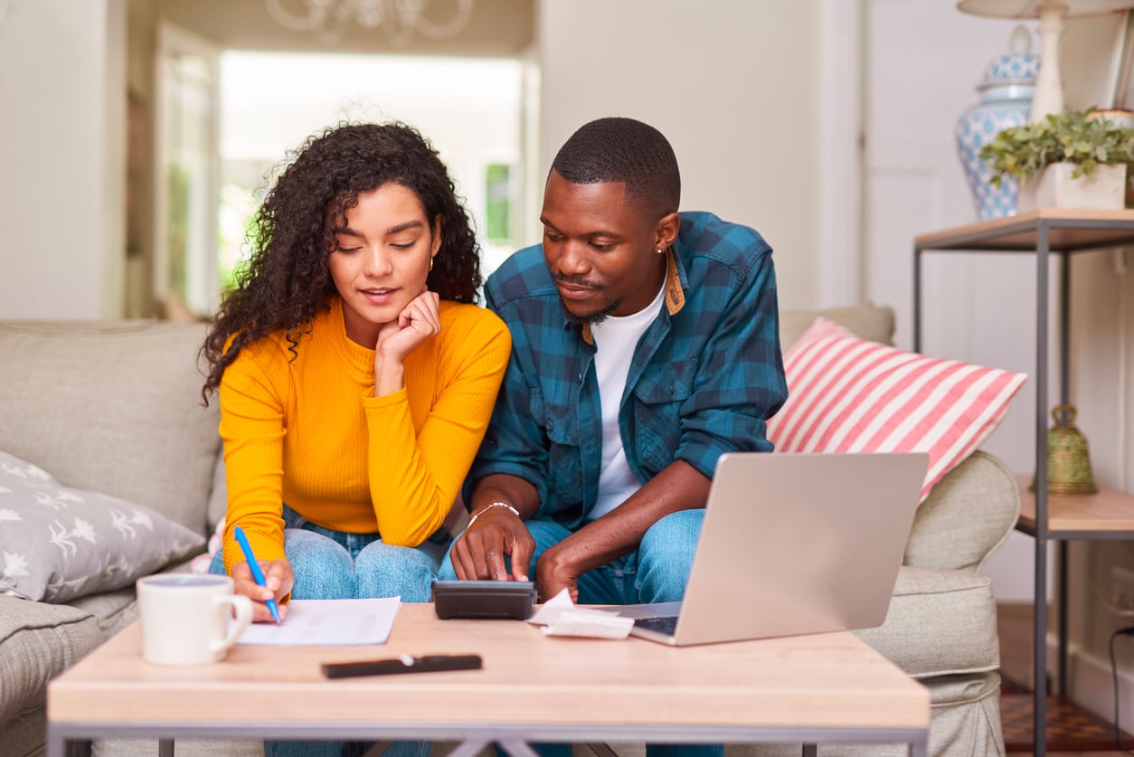 A couple sits on a couch, working together at a coffee table with papers, a pen, a calculator, and a laptop, appearing focused as they manage their household expenses in a cozy living room.