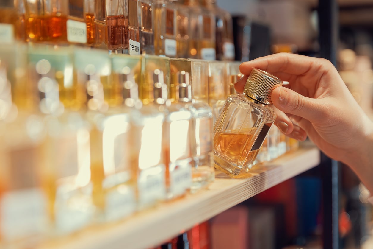 A hand selects a perfume bottle from a shelf lined with various glass fragrance bottles, all filled with different colored liquids.