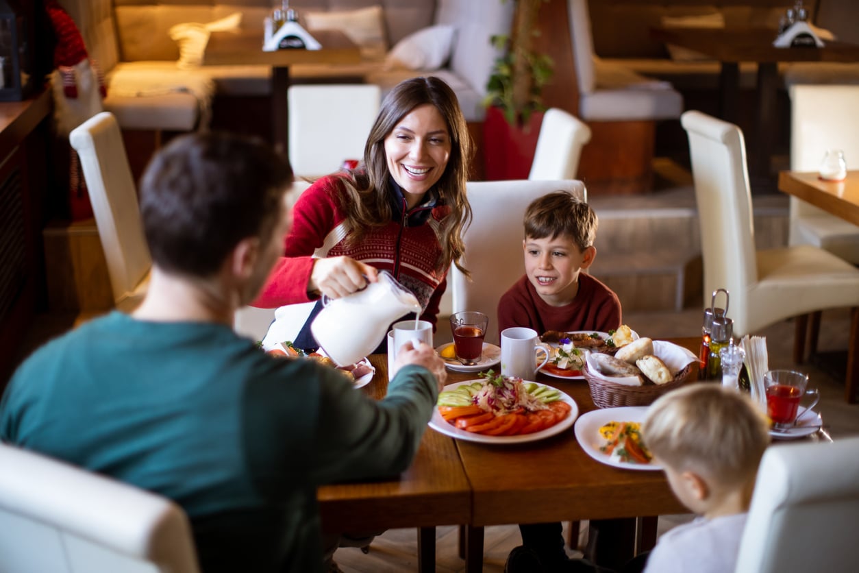 A woman pours a beverage for a man as two young boys sit at a restaurant table filled with food, including fruit, salad, and bread. The group appears to be enjoying a meal together, perhaps sharing stories from their recent vacation swaps.