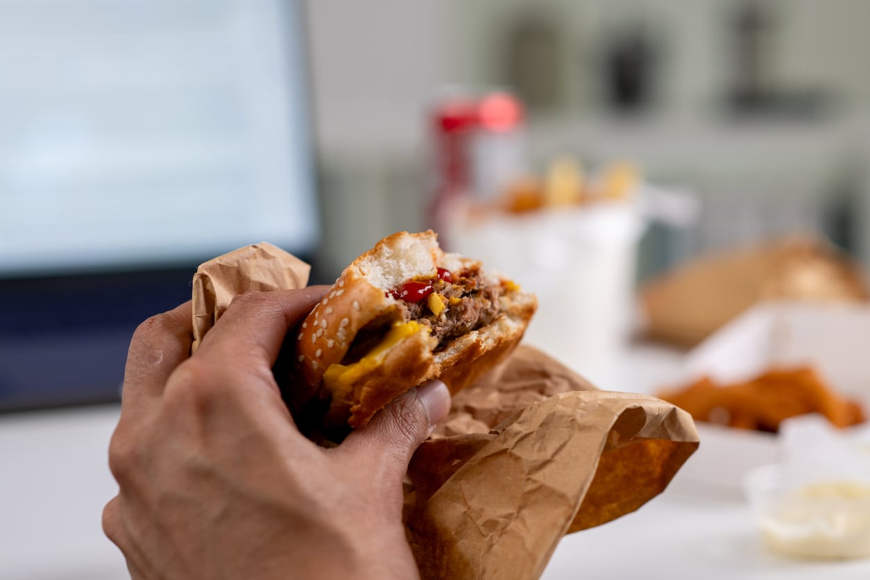 A person holding a half-eaten cheeseburger wrapped in brown paper, with a laptop, fries, chicken wings, and a soda can—some of the products people no longer buy as frequently—blurred in the background.