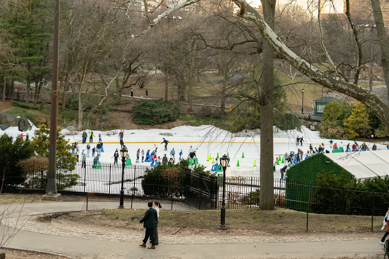 People ice skating on an outdoor rink in a park, surrounded by bare trees and patches of snow—a scene that captures the simple joys of winter or early spring. Two people walk along a path in the foreground, perhaps discussing which memberships are worth paying for to make the most of their time outdoors during the season.