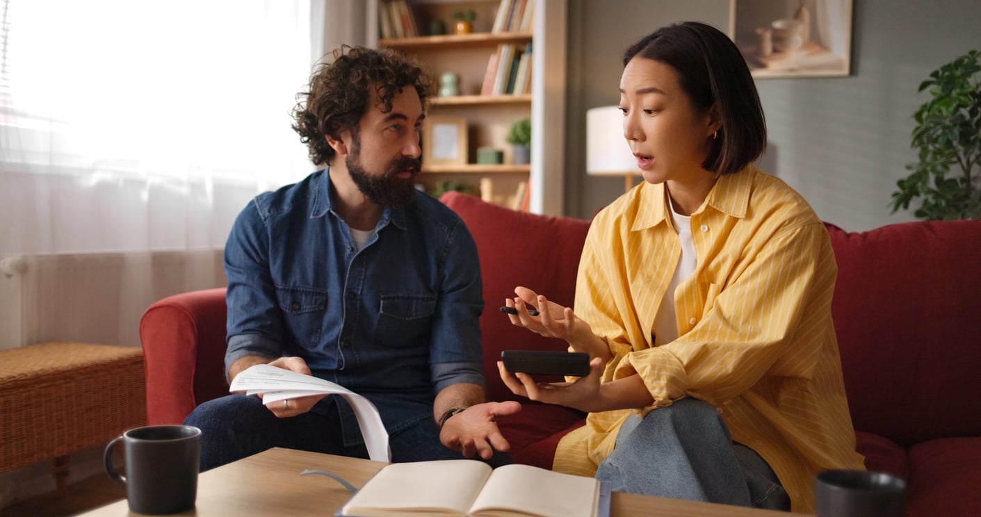 A man and woman sit on a red couch, talking seriously. The man holds papers, while the woman gestures and holds a calculator. An open notebook and two mugs are on the table in front of them.